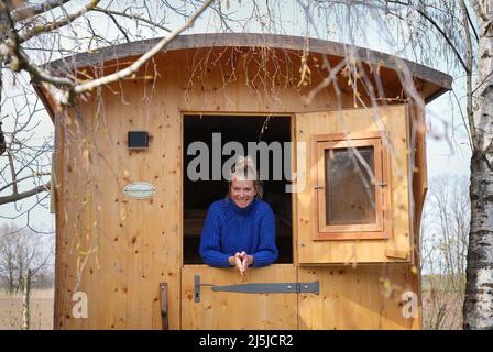 Kienitz, Deutschland. 12. April 2022. Die Berliner Urlauberin Claudia kleiner lehnt sich an den Erlenhof in der halb geöffneten Tür ihrer Schäferkarawane Schäfchenglück. Der Ferienbauernhof im Mittleren Oderbruch bietet naturnahe Ferien von April bis Oktober auf einer großzügigen Fläche und Übernachtungen in sechs Hirtenwägen sowie in einer Blockhütte. Maximal 15 Gäste finden auf dem Bauernhof Platz zum Schlafen, und es gibt weder Fernseher noch Radios in den Waggons und in der Kabine. (To dpa: ' 'Schäferstündchen im Schäferwagen' im Oderbruch') Quelle: Soeren Sache/dpa/Alamy Live News Stockfoto