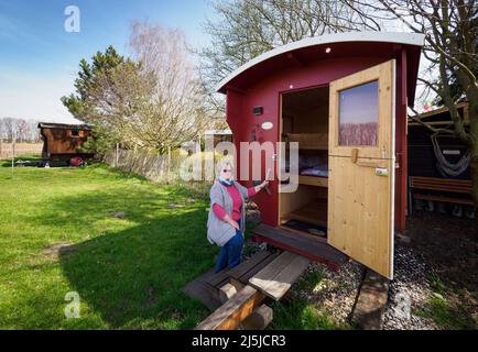 Kienitz, Deutschland. 12. April 2022. Barbara Brunat, Betreiberin des Erlenhofs, steht im Garten an der offenen Tür zum Hirtenwagen Wiesenschaf. Der Ferienbauernhof im Mittleren Oderbruch bietet naturnahe Ferien von April bis Oktober auf einem weitläufigen Gelände und Übernachtungen in sechs Hirtenwägen und in einer Blockhütte. Maximal 15 Gäste finden auf dem Bauernhof Platz zum Schlafen, und es gibt weder Fernseher noch Radios in den Waggons und in der Kabine. (To dpa: ' 'Schäferstündchen im Schäferwagen' im Oderbruch') Quelle: Soeren Sache/dpa/Alamy Live News Stockfoto