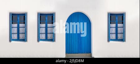 Holztür geschlossen und drei Glasfenster mit offenen Fensterläden blaue Farbe auf weißem Wandhintergrund. Kykladen Inselhaus Vorderansicht, griechisch traditionell Stockfoto