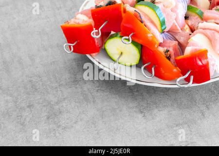 Roher putenfisch-Döner auf Metallspießen, mit viel buntem Gemüse, auf einem weißen Teller, bereit für die Zubereitung. Schreibtischoberfläche in hellgrauem Design Stockfoto