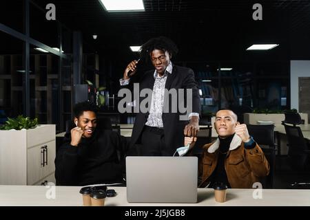 Multiethnische gemischte Rasse glückliche Kollegen mit Laptop. Schwarze afrikanische und asiatische Männer heben ihre Hände hoch, freuen sich über den Sieg und rufen, weil sie einen gewonnen haben Stockfoto