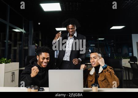 Multiethnische gemischte Rasse glückliche Kollegen mit Laptop. Schwarze afrikanische und asiatische Männer heben ihre Hände hoch, freuen sich über den Sieg und rufen, weil sie einen gewonnen haben Stockfoto