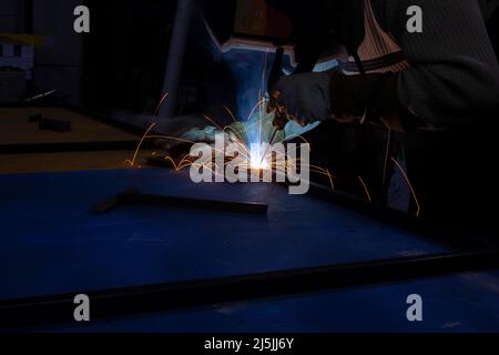 Schweißarbeiten. Ein Schweißer in einer Werkstatt schweißt ein Stahlstück. Garagenarbeit für Männer. Funken durch Schweißen. Arbeiter trägt eine Maske, um seine Augen vor Feuer zu schützen. Stockfoto