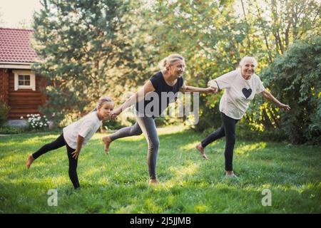 Drei Generationen von Familienfrauen, die lächelnd auf einem Bein balancieren, halten die Hände zusammen, um auf einem grünen Rasen auf dem Hinterhof körperliche Übungen zu machen Stockfoto