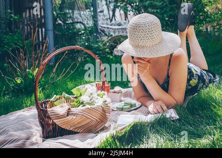 Junge Frau, die auf Karli liegt und im Garten auf Gras ein Buch liest. Psychische Gesundheit, Wellness gesundes Lifestyle-Konzept. Picknick im Sommer Stockfoto