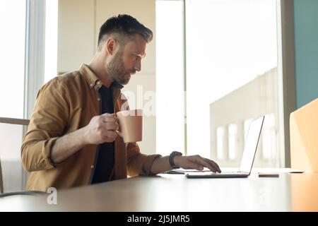 Mann, Der Am Laptop Arbeitet Und Kaffee Im Büro Trinkt, Seitenansicht Stockfoto
