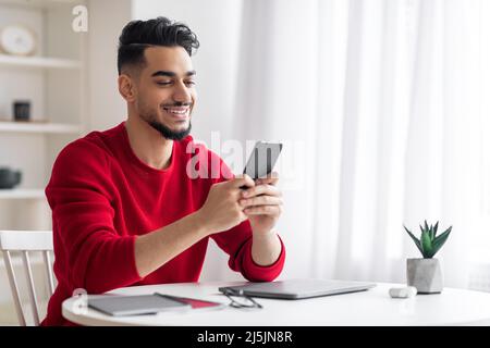 Froher attraktiver junger arabischer Mann mit Bart in roter Kleidung, der am Arbeitsplatz eine Nachricht auf dem Smartphone tippt Stockfoto