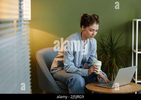 Porträt einer jungen Frau in Freizeitkleidung mit Laptop an der Ecke des Arbeitsplatzes gegen die grüne Wand Stockfoto