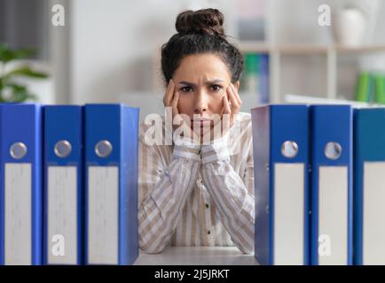 Verärgerte Geschäftsfrau, die mit einem Stapel von Ordnern am Arbeitsplatz sitzt und die Kamera anschaut und sich müde fühlt Stockfoto
