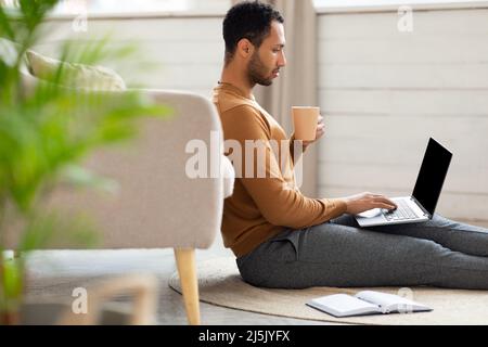 Fokussierter arabischer Mann, der sich ein Video am Computer ansieht und Kaffee trinkt Stockfoto