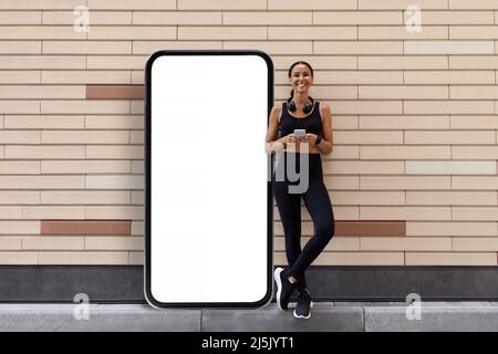 Lächelnde junge arabische Sportlerin steht in der Nähe des riesigen Smartphones mit leerem Bildschirm Stockfoto