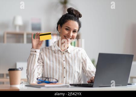 Porträt einer glücklichen Geschäftsfrau, die ihre Kreditkarte hält, während sie auf einem Laptop online einkaufen und vor der Kamera lächelt Stockfoto
