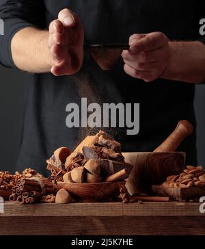 Der Koch streut die Schokolade mit Kakaopulver. Auf einem alten Holztisch Süßigkeiten, Kaffeebohnen, Zimt, Anis, Nüsse, und Stücke von gebrochenem schwarzem Choco Stockfoto