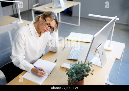 Müde Büroangestellte, die am Arbeitsplatz in einem Notizbuch schrieb Stockfoto