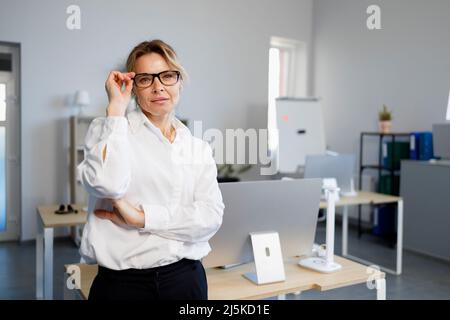 Selbstbewusste reife Geschäftsfrau in Brille und weißem Hemd steht im Büro Stockfoto