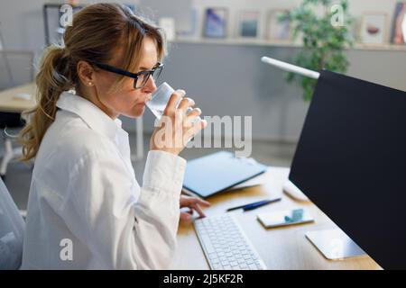 Seitenansicht einer Geschäftsfrau mittleren Alters, die Wasser trinkt und am Computer sitzt Stockfoto
