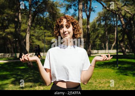 Unsichere Frau, die weiße T-Shirts und schwarze Yogahosen trägt, die im Freien mit den Schultern zucken. Junge Rotschopf-Frau schaut weg. Zögernde Frau. Hilflos Stockfoto