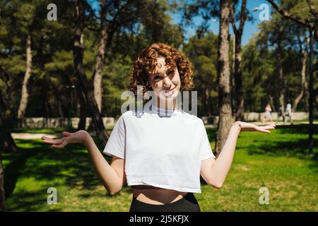 Unsichere Frau, die weiße T-Shirts und schwarze Yogahosen trägt, die im Freien mit den Schultern zucken. Attraktive Rothaarige Frau mit Blick auf die Kamera. Zögernde Frau Stockfoto