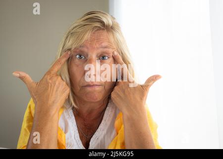 Kaukasische Frau, 50-60 Jahre alt, mit blondem Haar, das mit beiden Händen auf den Kopf zeigt. Burnout-Konzept. Stockfoto