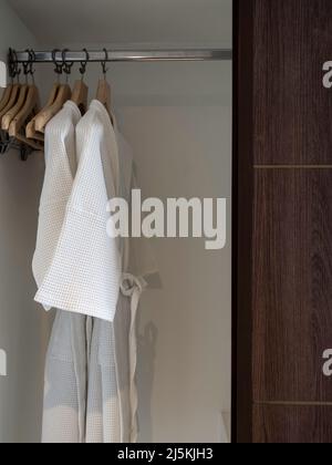 Ein paar weiße, saubere Bademäntel hängen an den Holzbügeln im Holzschrank für die Hotelgäste im Zimmer, vertikaler Stil. Stockfoto