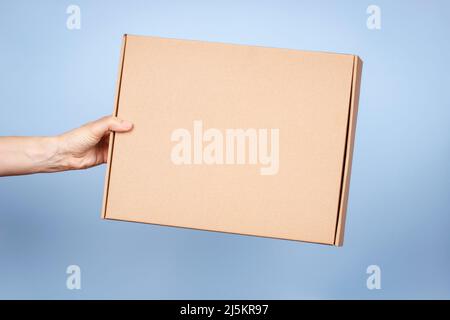 Frau Hand hält braunen ökologischen Verpackungskarton aus natürlicher Wellpappe. Mockup-Versandkarton auf blauem Hintergrund. Verpackung Stockfoto