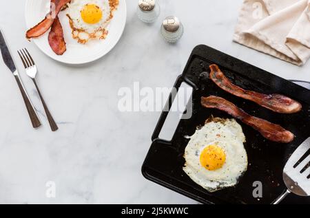 Blick von oben auf Speck und Eier auf einer Grillpfanne und auf einem Teller zum Frühstück. Stockfoto