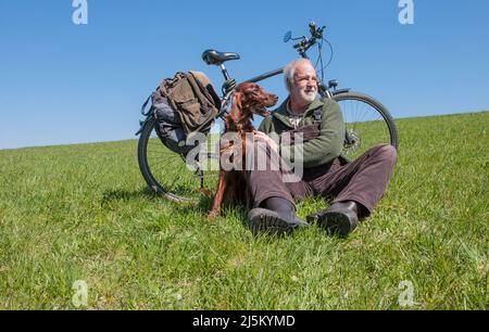 Ein älterer mit einem Fahrrad sitzt auf einer grünen Wiese im Sonnenschein und hält seinen irischen Setter-Jagdhund liebevoll in den Armen. Stockfoto