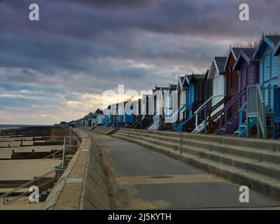 Farbenfrohe Strandhütten, eine Esplanade und ein groyner Strand in einem jenseitigen, schrägen Licht von einer untergehenden Sonne, verdeckt von stürmischen Wolken. Stockfoto