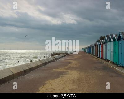 Bunte Strandhütten blicken über die sonnenbeschienen Nordsee-Gewässer, während eine Möwe Patrouille über sich legt. Ein gespenstiger Windpark ist nur am Horizont sichtbar. Stockfoto