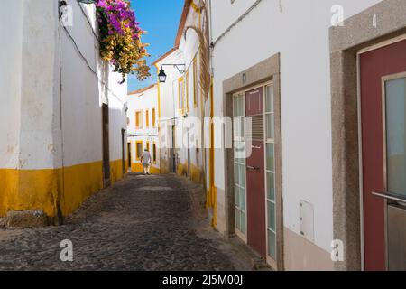 Älterer Mann, der in einer der malerischen Gassen mit Kopfsteinpflaster mit weiß getünchten und gelben Häusern im historischen Zentrum von Evora, Portugal, spaziert. Stockfoto