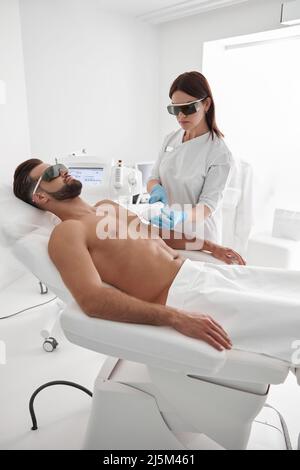 Ein junger Meister mit Brille führt eine Laserepilation bei einem bärtigen Patienten durch, der in der Klinik auf der Couch liegt Stockfoto