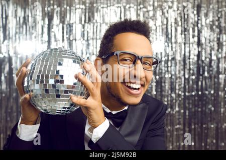 Glücklicher junger afroamerikanischer Mann in Anzug und Brille, hält einen Diskoball und lächelt Stockfoto