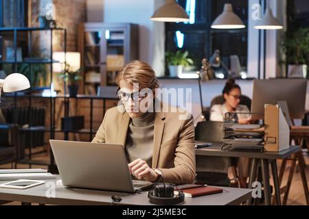 Junger kaukasischer Mann mit Brille und Jacke, der am Schreibtisch sitzt und einen Laptop benutzt, während er im Dark Office arbeitet Stockfoto