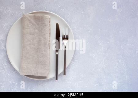 Weißer runder Teller mit Besteck und Leinenserviette auf hellgrauem Betongrund, Minimalismus-Stil, Draufsicht, Kopierraum, Stockfoto