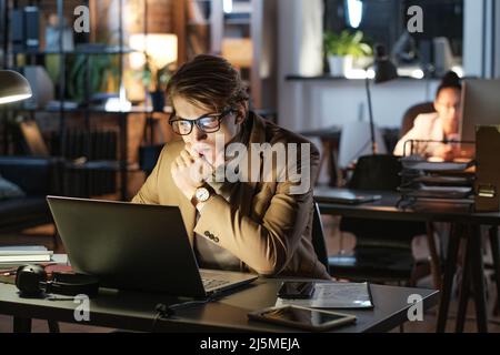 Fokussierter junger Geschäftsmann in einer Brille, der das Kinn berührt und im dunklen Büro mit einem Laptop arbeitet Stockfoto