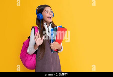 Erstauntes Kind in Kopfhörern mit Schulrucksack mit Arbeitsbuch auf gelbem Hintergrund Stockfoto