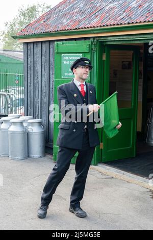 Wache in traditioneller Uniform der British Railways, pfeifend und mit grüner Flagge winkend. Watercress Railway Line, Hampshire, England Stockfoto