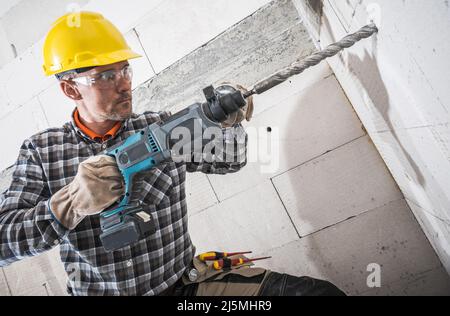 Kaukasischer Bauarbeiter in seinem 40s mit leistungsstarkem Schlagbohrer. Bohren notwendig ein Loch in Beton Ziegelwand. Stockfoto