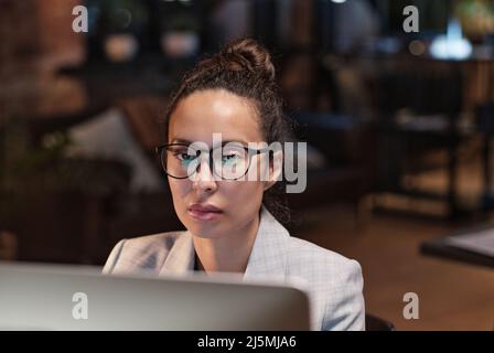 Beschäftigte junge hispanische Frau mit Brille, die im dunklen Büro sitzt und mit dem Computer arbeitet, während sie ihre Aufgabe fristgerecht erledigt Stockfoto