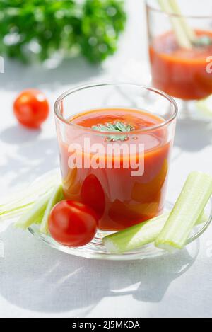 Frischer Tomatensaft. Tomatensaft wird in ein transparentes Glas mit Petersilie und Sellerie-Stielen gegossen, auf weißem Hintergrund, beleuchtet durch Sonnenlicht Stockfoto