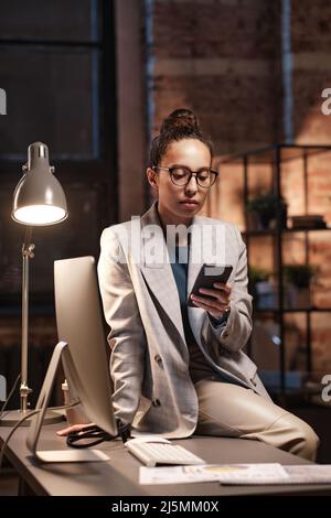 Seriöse junge Geschäftsfrau mit gemischter Rasse in Brillen, die mit Computer auf dem Tisch sitzt und im dunklen Büro durch soziale Medien scrollt Stockfoto