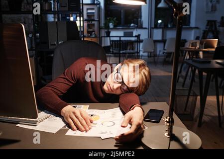 Müde, junger kaukasischer Manager mit Brille, der am Schreibtisch saß und in einem leeren dunklen Büro auf Geschäftsunterlagen schlief Stockfoto