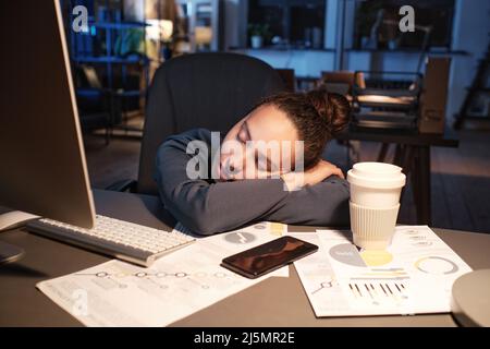 Müde junge Frau mit gemischter Rasse, die sich auf gekreuzte Arme lehnte, während sie am Arbeitsplatz im dunklen Büro schlief Stockfoto
