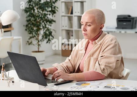 Seriöse junge kahle Geschäftsfrau mit Rollkragen und Hemd, die im Büro am Schreibtisch sitzt und mit einem Laptop arbeitet Stockfoto