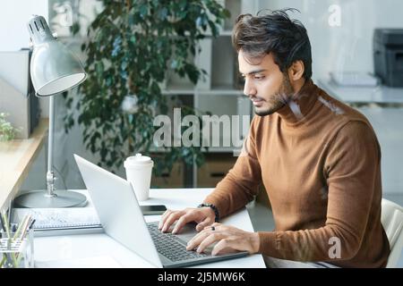 Konzentrierter junger Mann aus dem Nahen Osten in braunem Rollkragen, der am Tisch sitzt und im Büro einen Laptop benutzt Stockfoto