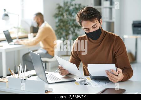 Beschäftigter junger gemischter Büromann mit schwarzer Stoffmaske, der mit einem Laptop am Tisch sitzt und den Bericht prüft Stockfoto