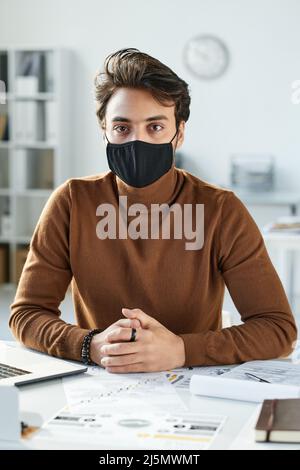 Porträt eines jungen Mannes aus dem Nahen Osten in schwarzer Maske, der mit Papieren am Tisch sitzt und an einem Bericht arbeitet Stockfoto