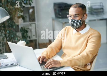Konzentrierter junger multiethnischer Account Manager in Gesichtsmaske und Brille mit Laptop, während er Internetforschung analysierte Stockfoto