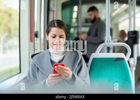 Positive Frau liest vom Handy-Bildschirm in der Straßenbahn Stockfoto