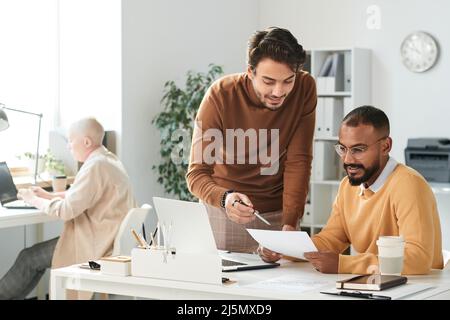 Gutaussehende junge Führungskraft zeigt auf Papier, während sie mit einem Kollegen im Büro über die Projektstrategie spricht Stockfoto